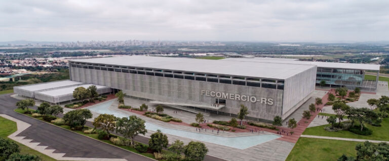 Fachada extensa de edifício comercial sustentável com painéis de sombreamento perfurados, estrutura suspensa por pilares sobre um espelho d'água e pedestres circulando em um amplo deck de madeira.